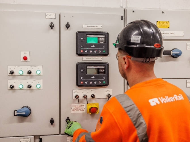 A man standing and looking at a high voltage power unit wearing a black hard hat and orange PPE